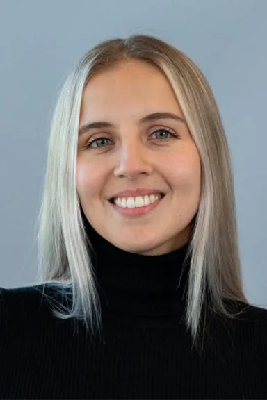 Ksenia Moskalenko in a black turtleneck against a grey-blue background, smiling